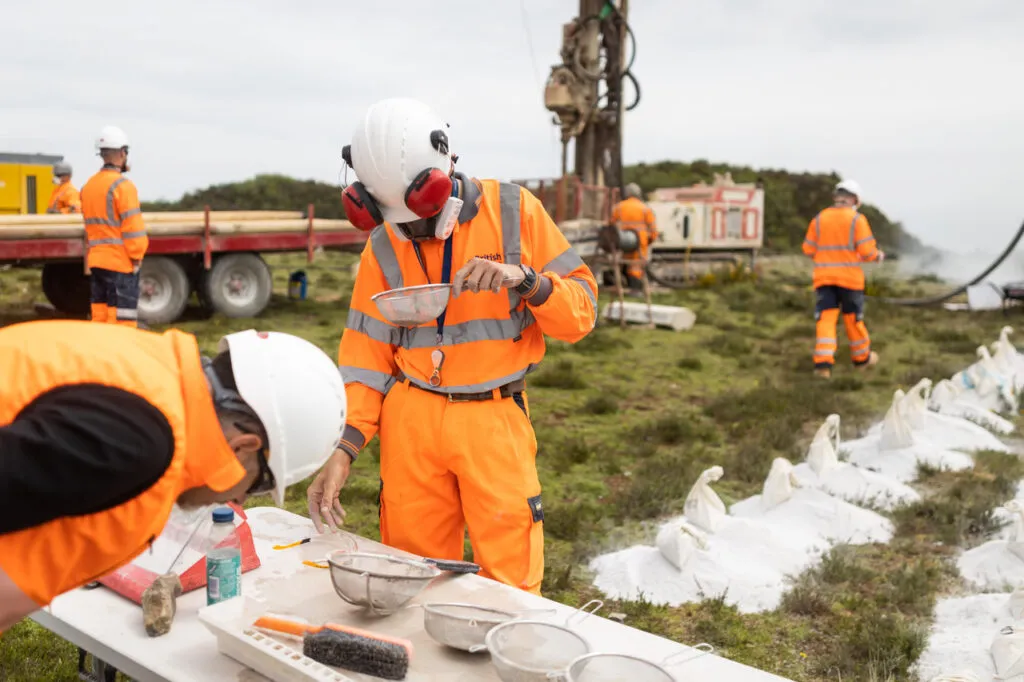 Geologists in high-visibility gear examining lithium samples during fieldwork at a drilling site with equipment and sample bags.