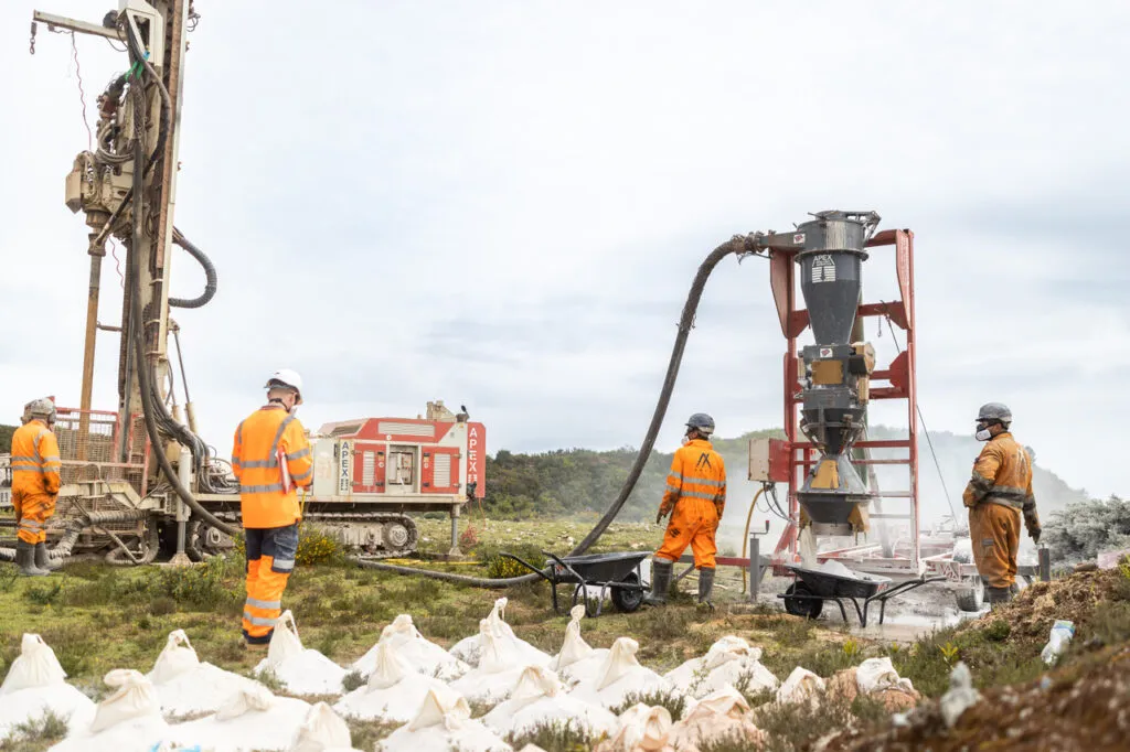 Workers operating drilling equipment at a lithium exploration site in Cornwall, wearing safety gear and surrounded by machinery and sample bags.