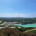 Aerial view of an open-pit quarry with turquoise water, green hills and winding roads under a blue sky in Cornwall.