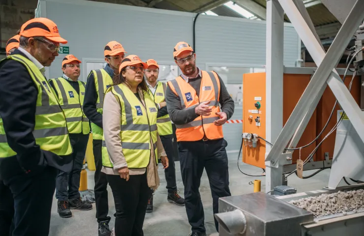 Group of visitors in safety gear touring British Lithium facility, observing equipment and listening to a technical explanation.