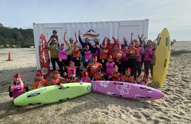 Group of children and instructors in surf gear posing on the beach with rescue boards, celebrating surf lifesaving training.