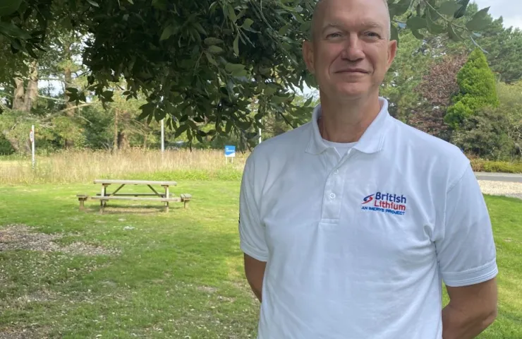 Man wearing British Lithium polo shirt standing outdoors in a green park area with trees and a picnic bench behind.