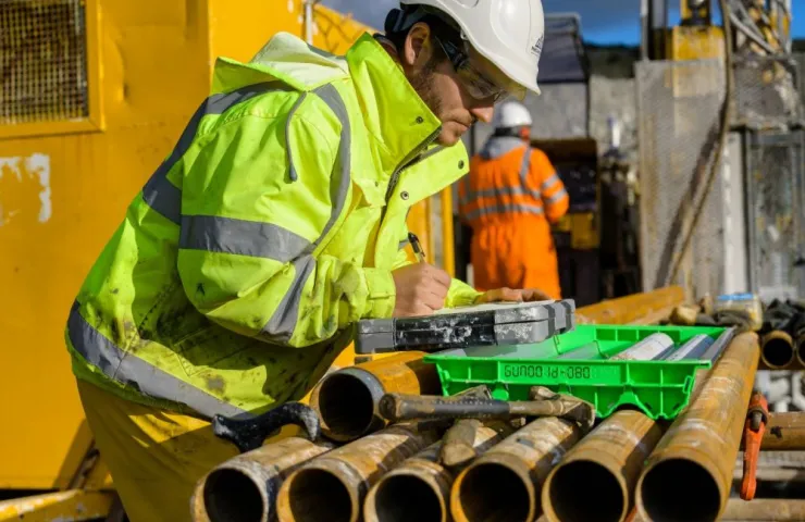 Geologist in safety gear marking core samples at a lithium exploration drill site with pipes and equipment in background.